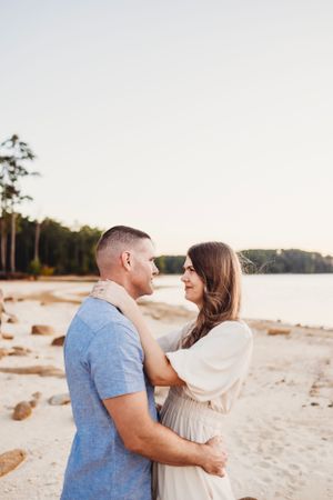 A romantic couple embraces on a sandy beach at sunset with tall pine trees in the background.