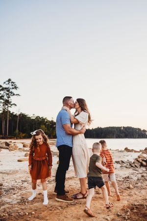 Family enjoying a tender moment together on a scenic beach with pine trees and water in the background.