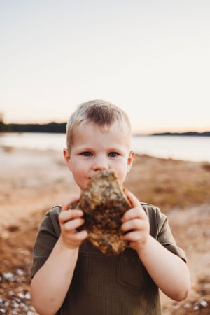 Young child holding a large rock while standing on a beach at sunset.