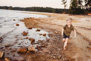 Child walking along rocky shoreline at sunset with lake and trees in background.