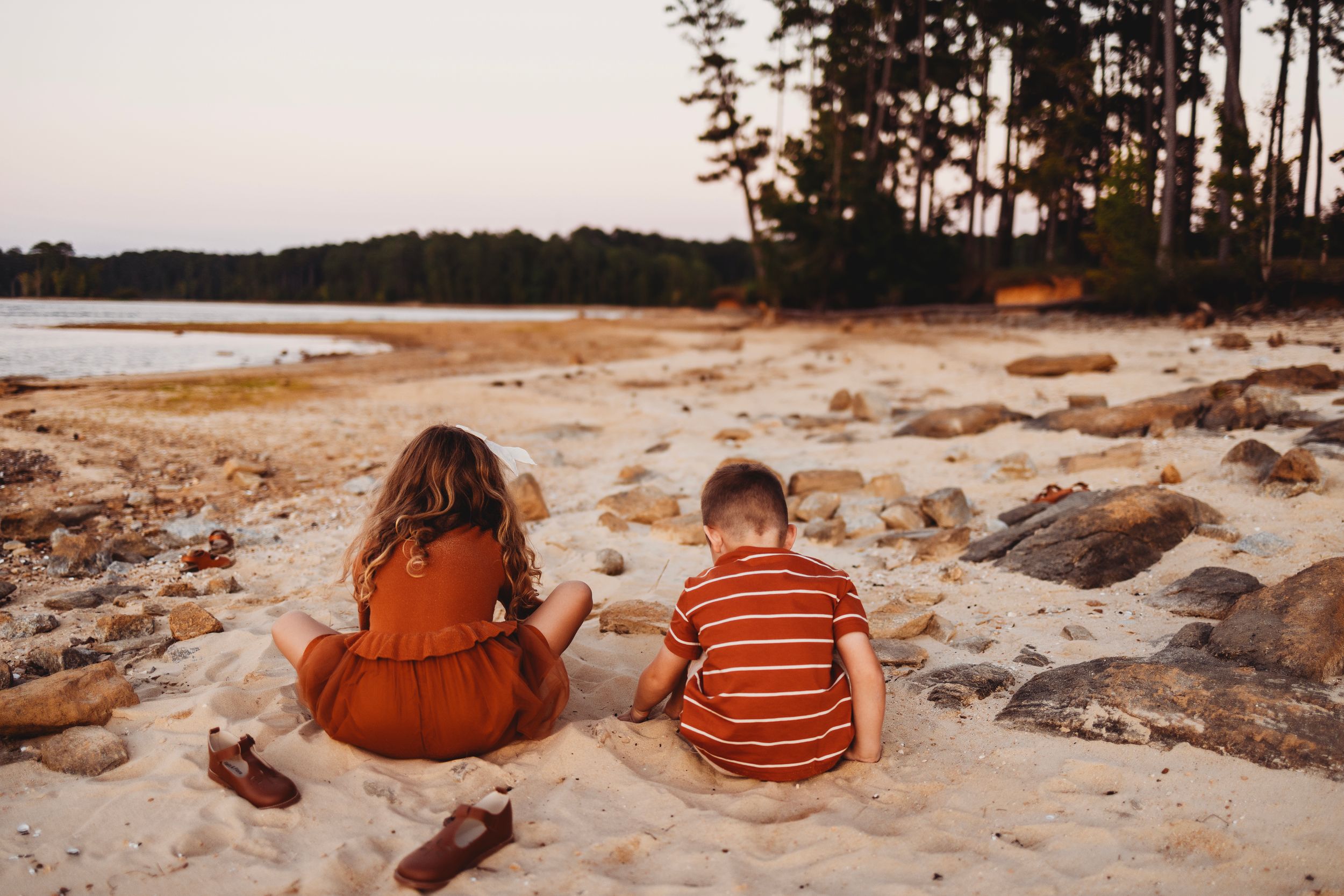 Two children sitting together on sandy beach looking at water during sunset.