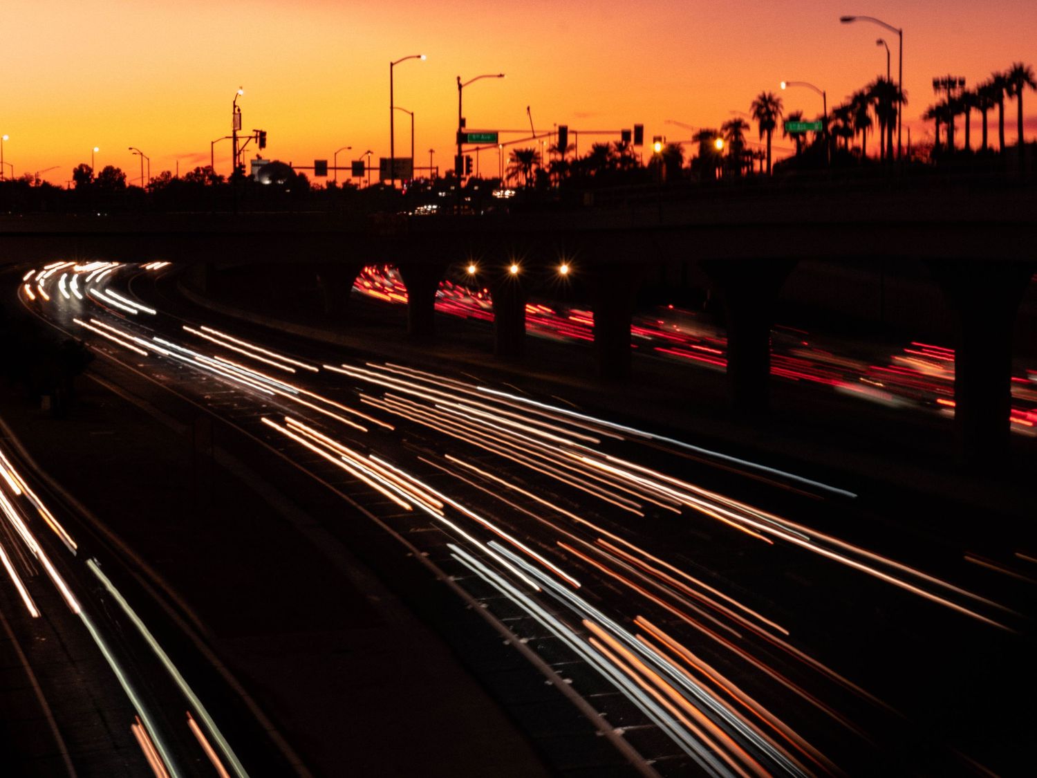 Long exposure of car lights streaking along highway at sunset with palm trees silhouetted against Phoenix orange sky.