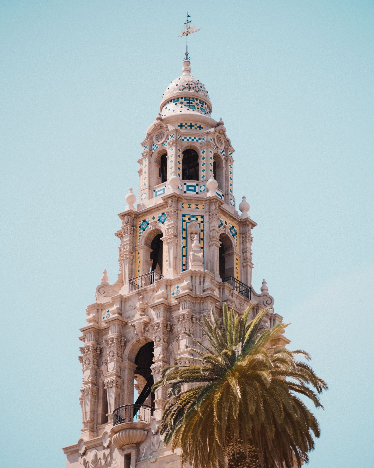 Ornate Spanish colonial church tower with intricate details and a palm tree against clear blue sky.