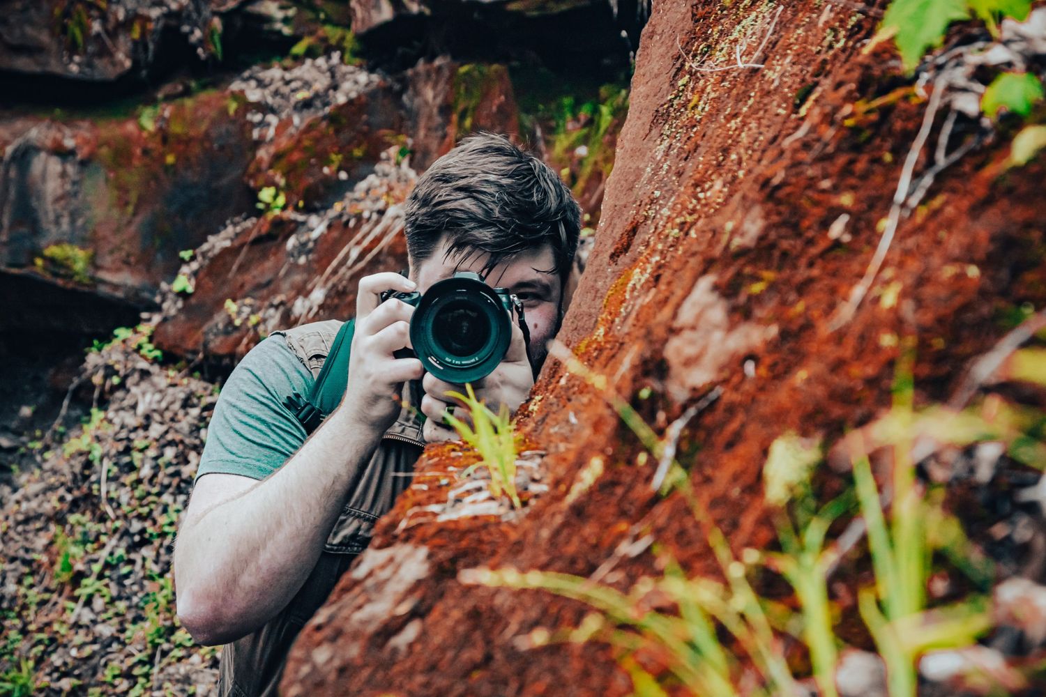 A photographer with a camera captures a photo against a rustic red rock wall with green foliage.