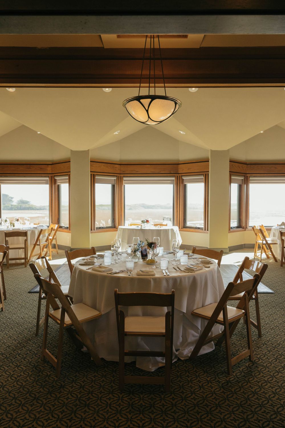 Elegant round dining table set for a wedding reception with wooden chairs and panoramic windows overlooking scenic views.