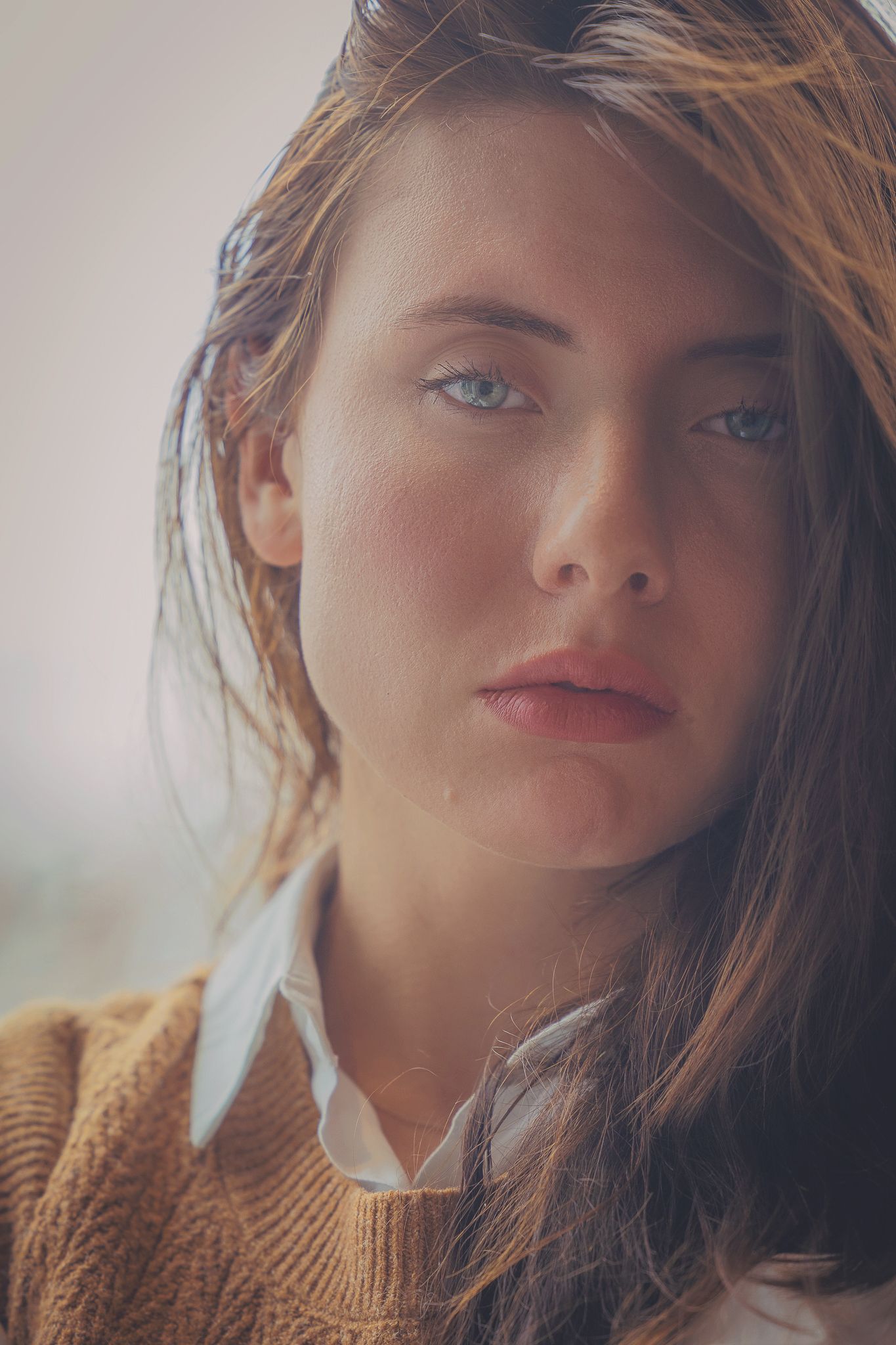 Artistic portrait in warm lighting shows piercing blue eyes and brown sweater with tousled brown hair falling naturally.