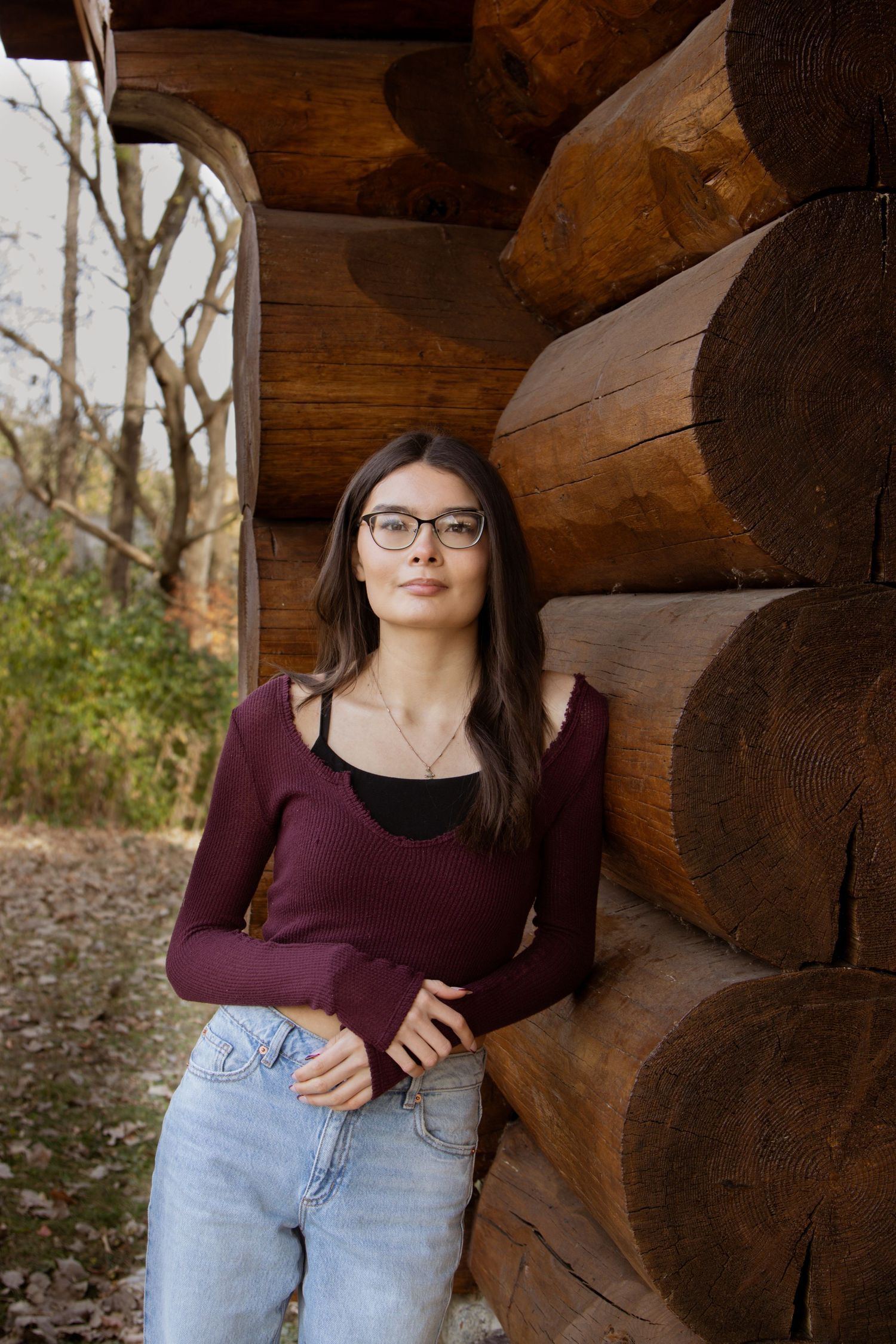 Young woman in burgundy top and jeans poses by rustic log cabin wall.