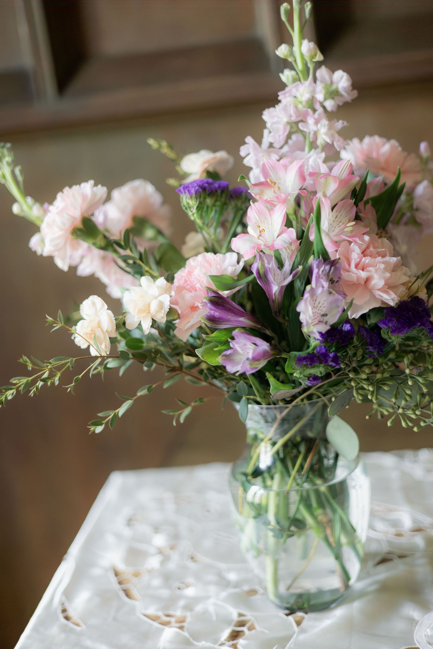 A mason jar vase filled with pink carnations, purple flowers, and greenery on a lace tablecloth.