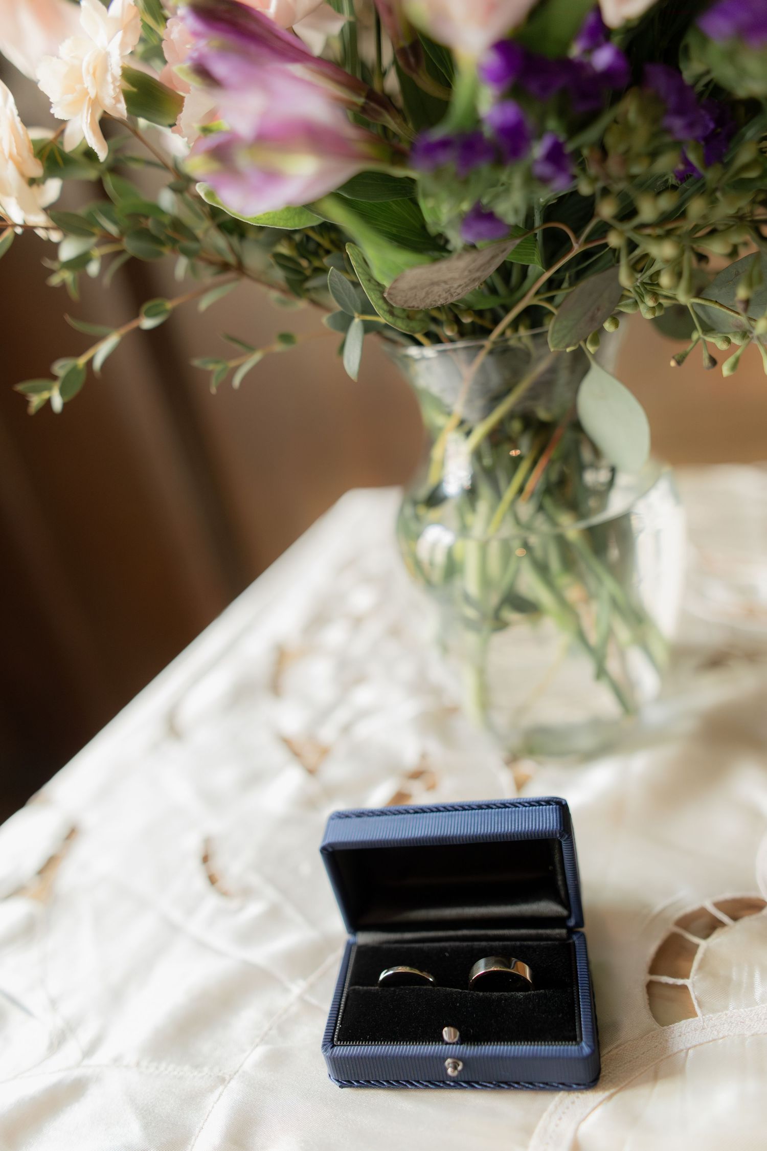 Wedding rings in a navy blue box next to floral centerpiece on white tablecloth.