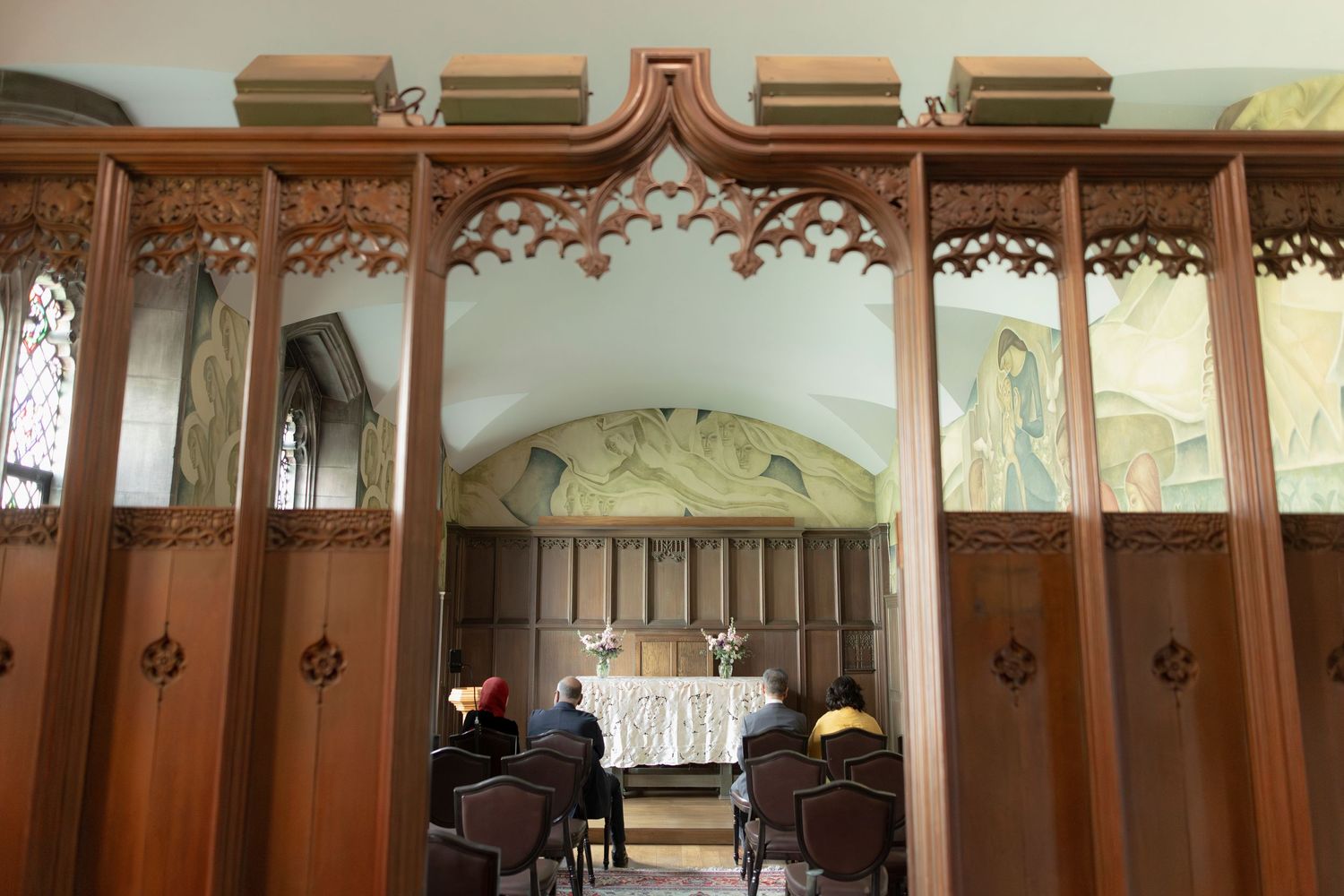 A view through decorative wooden archways into a small chapel with altar and chairs.