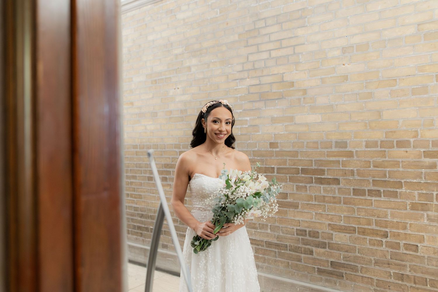 A bridal portrait featuring a white wedding dress and delicate bouquet against a brick wall background.