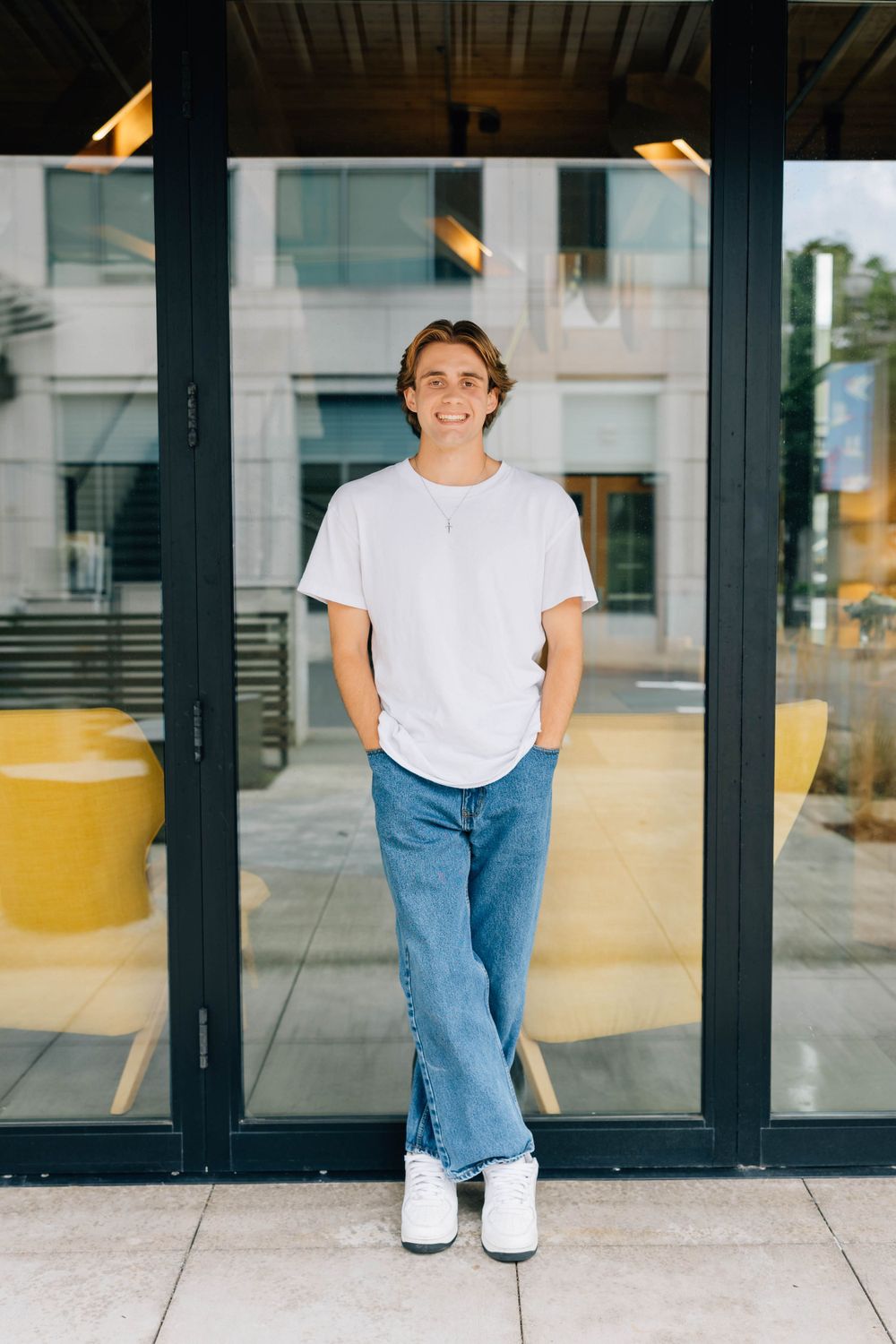 Smiling young man in white t-shirt and jeans standing confidently by glass doors outdoors.