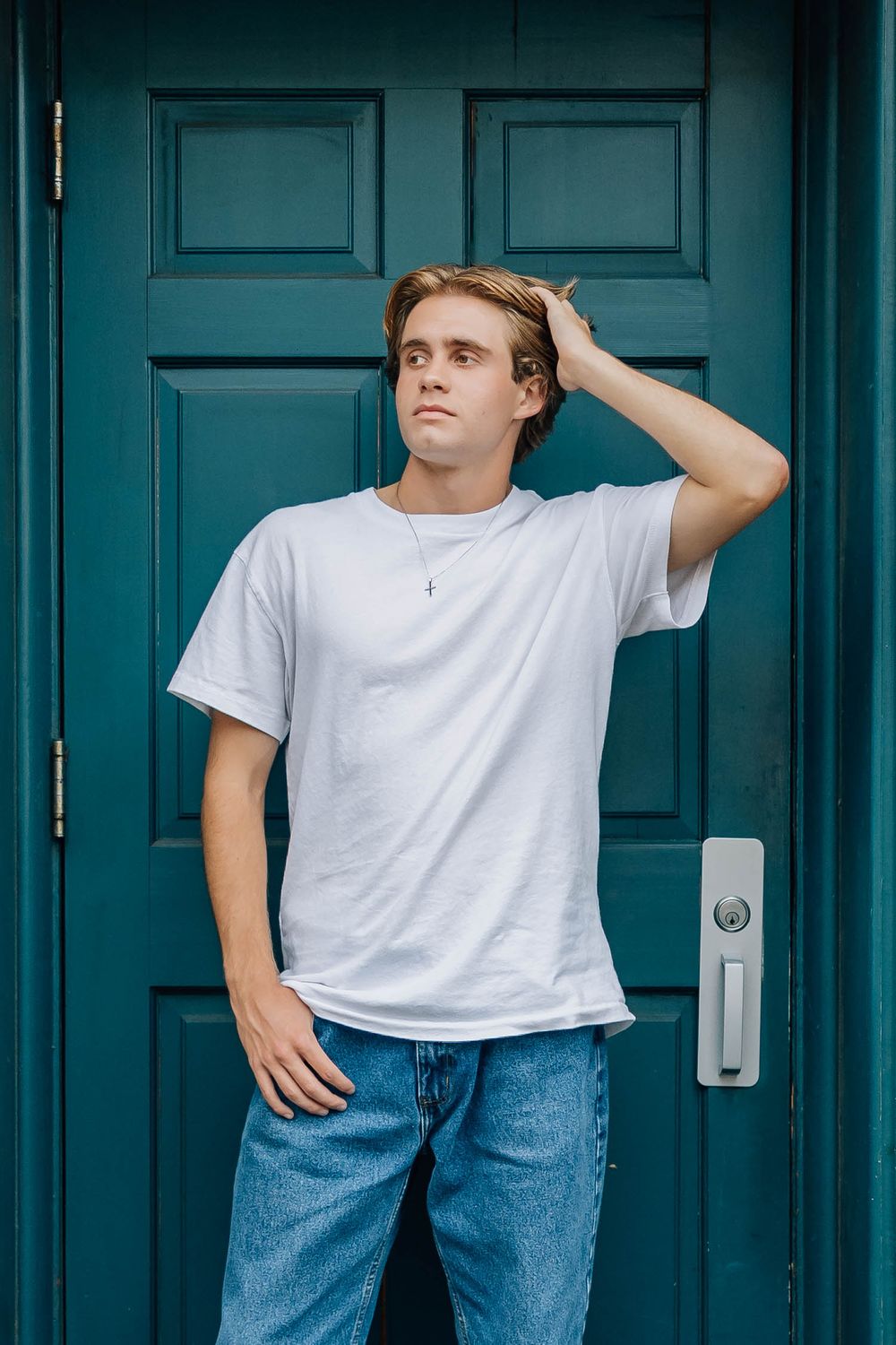 Young man in white t-shirt running hand through hair while standing before a teal door.