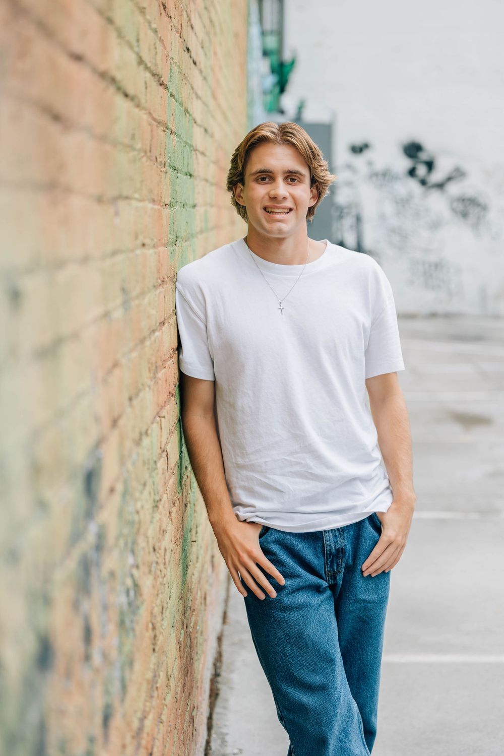 Smiling young man in white t-shirt and jeans leaning casually against a brick wall outdoors.