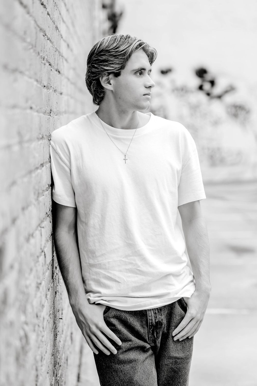 Young man in white t-shirt and jeans leaning against brick wall, black and white portrait photo.