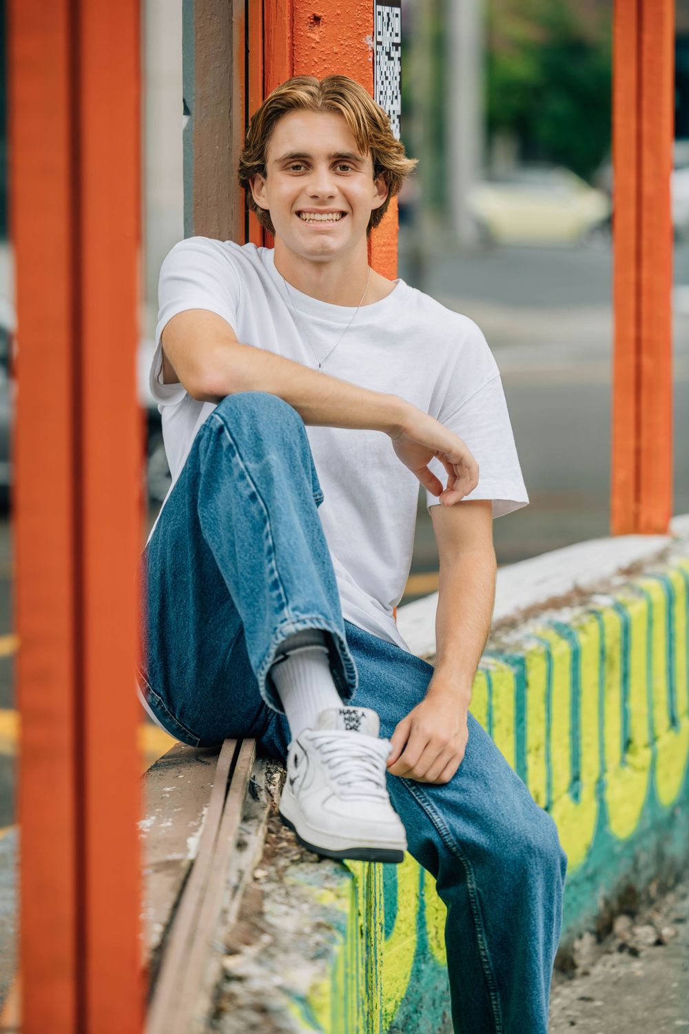 Cheerful young man in white tee and jeans sitting relaxed against orange pillar with graffiti.