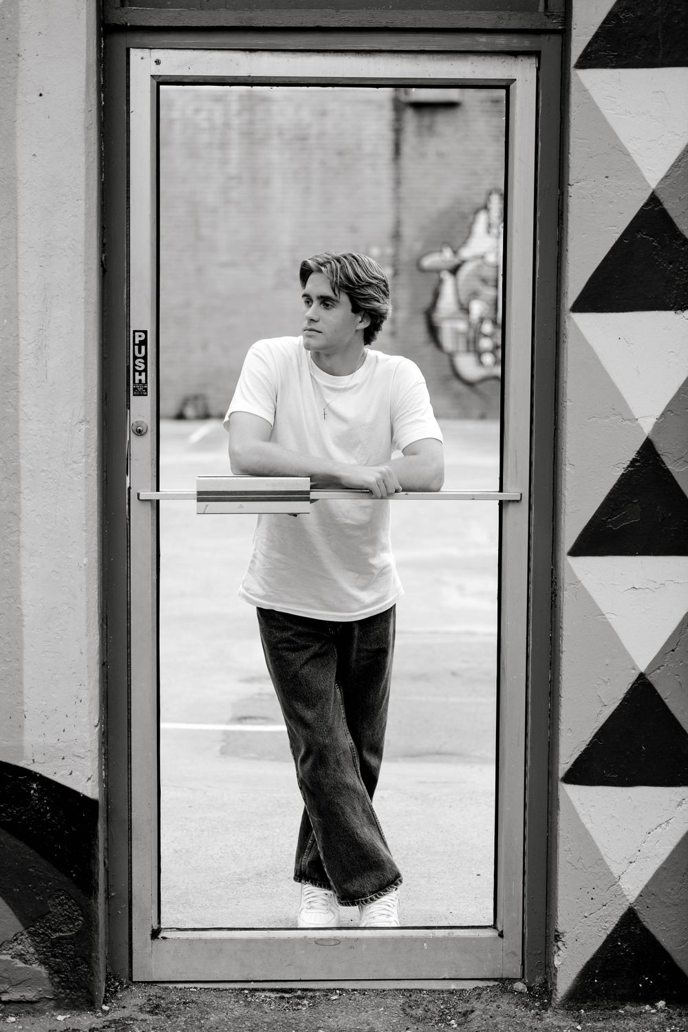 Young man in white t-shirt leaning on glass door handle, black and white urban portrait.