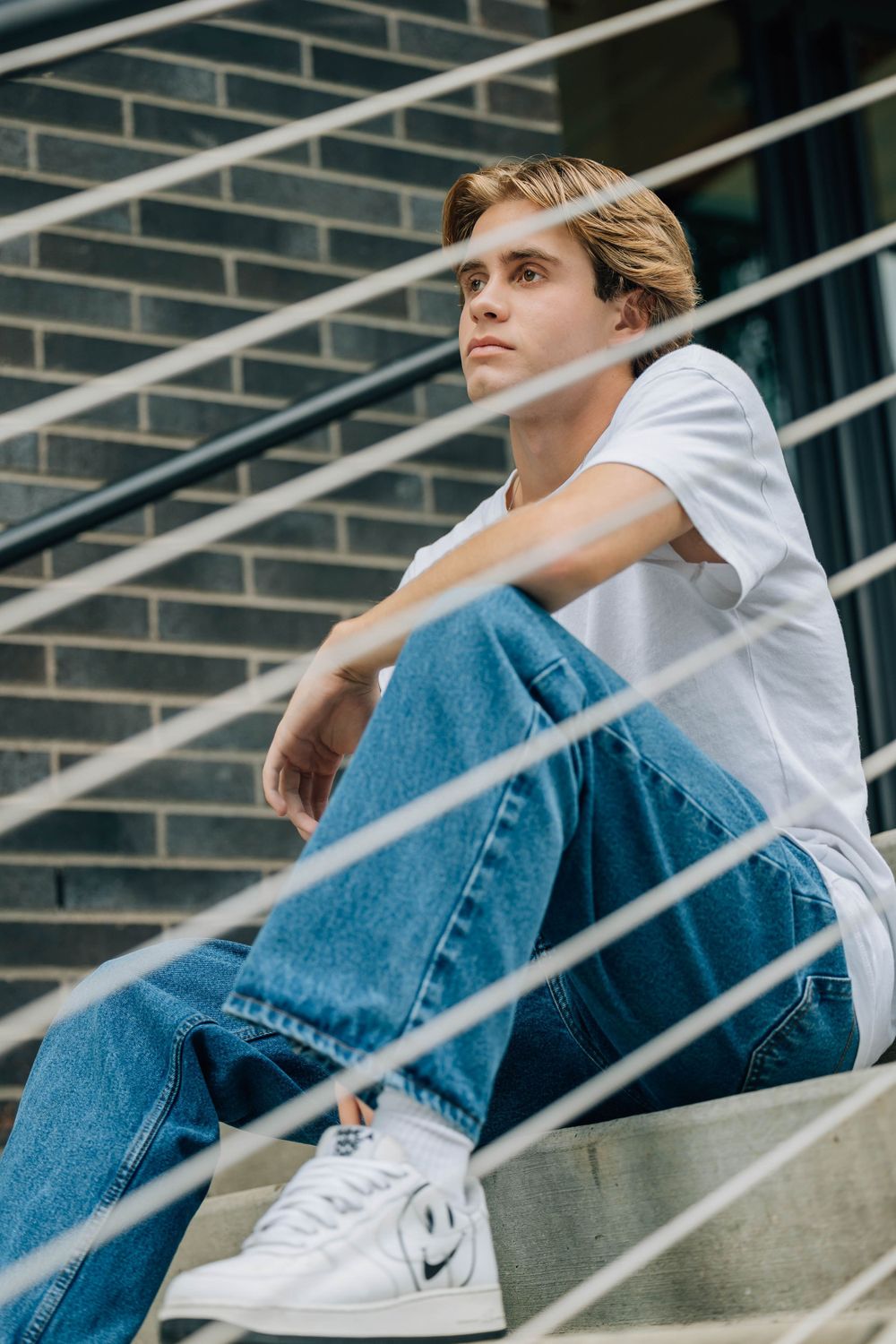 Young man in white tee and blue jeans sitting on urban steps with metal railing.