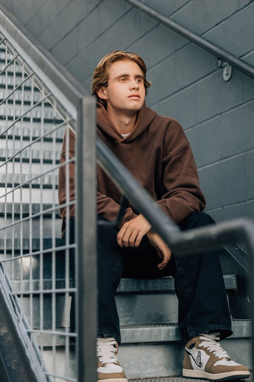 Young man in brown hoodie sitting pensively on metal stairs with grid railing in urban setting.