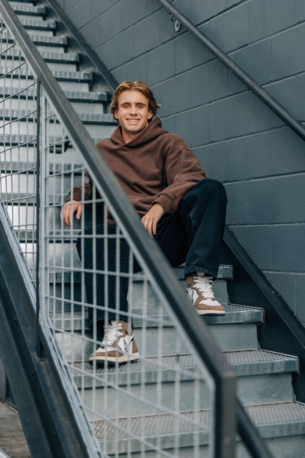 Smiling young man in brown hoodie sitting on industrial metal staircase against gray wall.