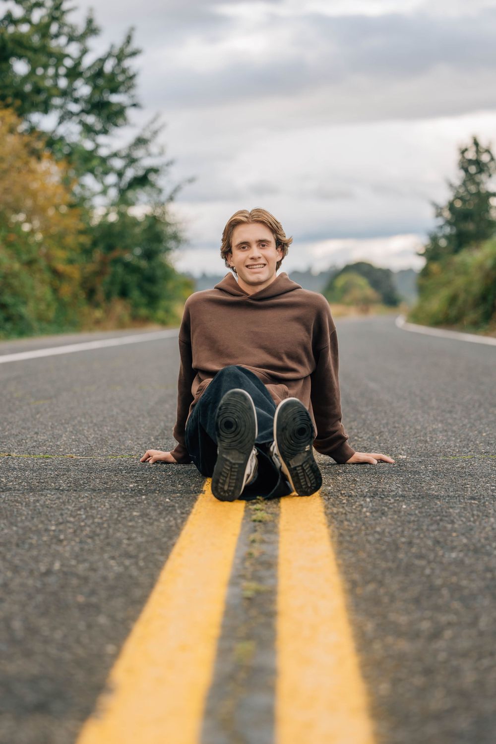 Smiling teenage boy in brown hoodie sitting on yellow center line of empty road.