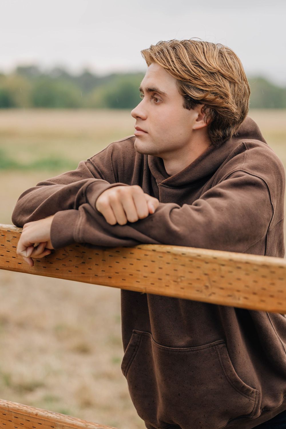 Thoughtful young man in brown hoodie leaning on wooden fence rail in open field.