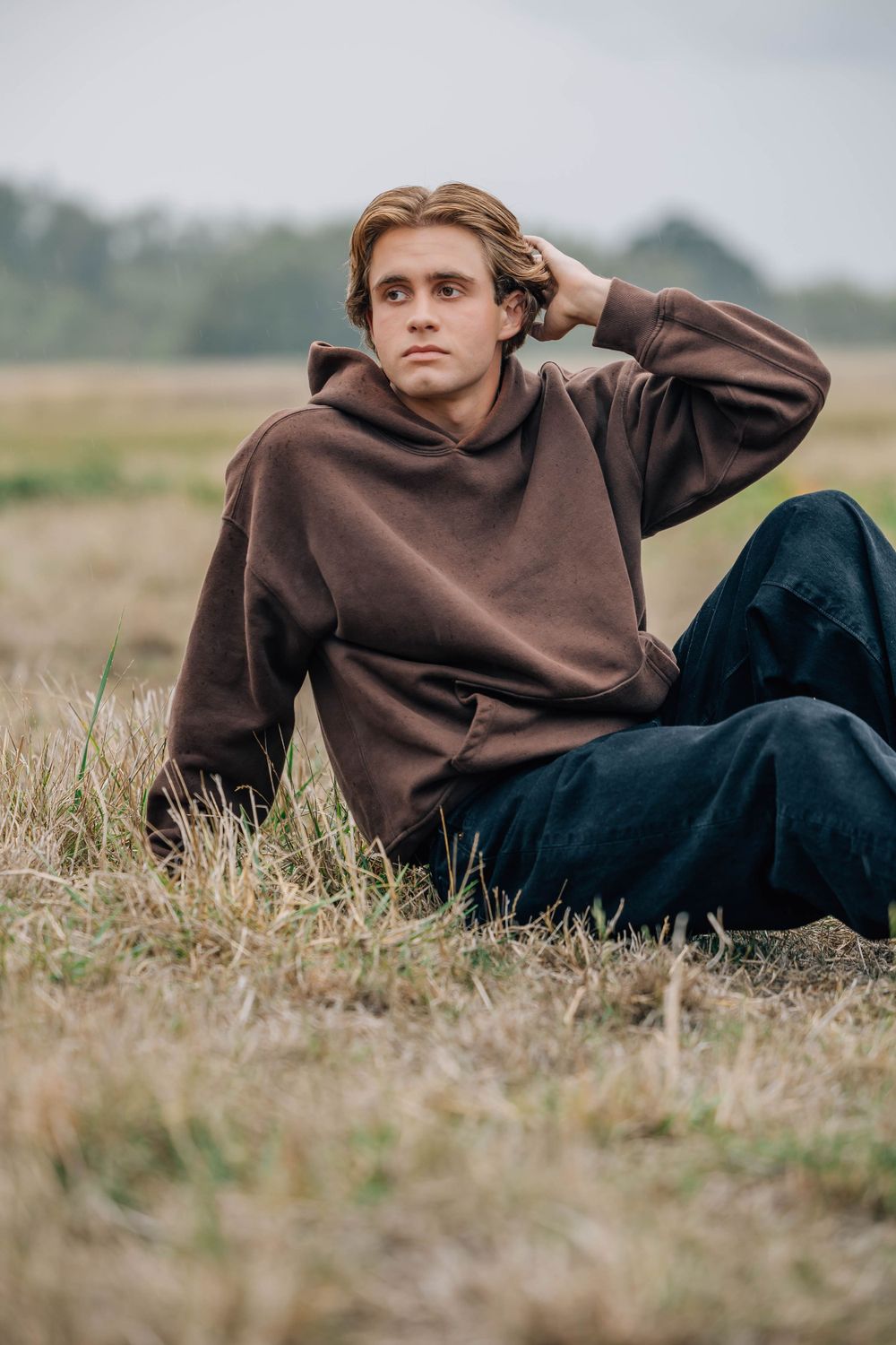 Young man in brown hoodie sitting in dry grass field with hand in hair.