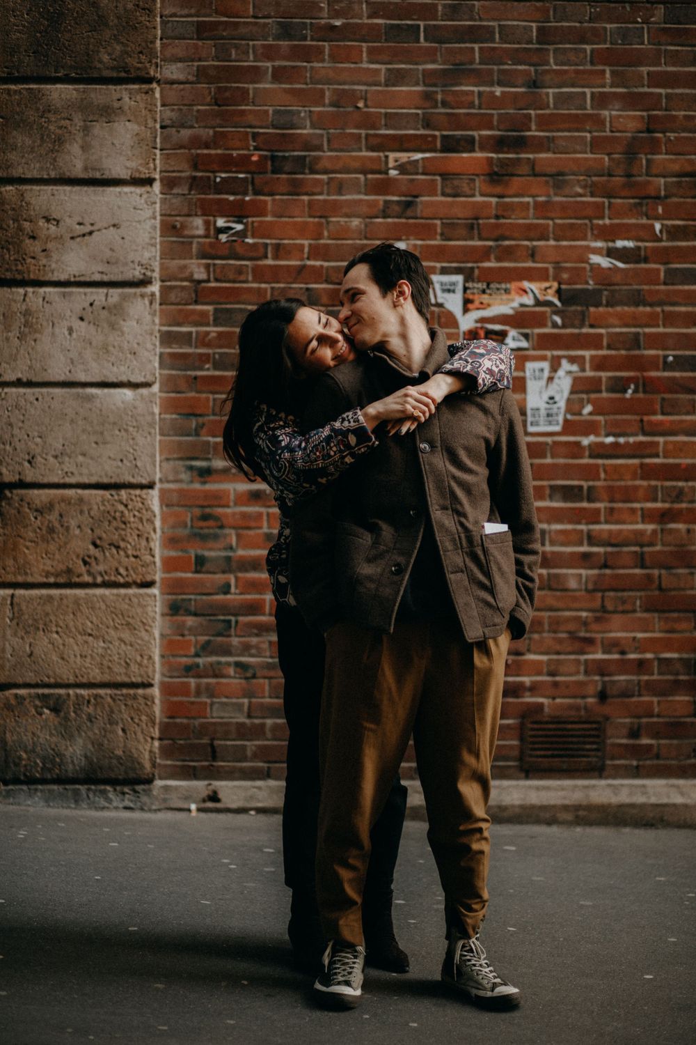 A couple laughing and embracing against an urban brick wall background.