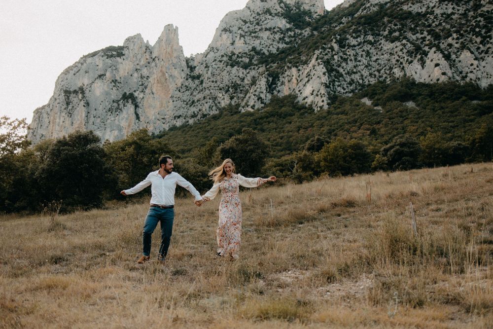 Two people running joyfully through a field with dramatic mountain cliffs in the background.