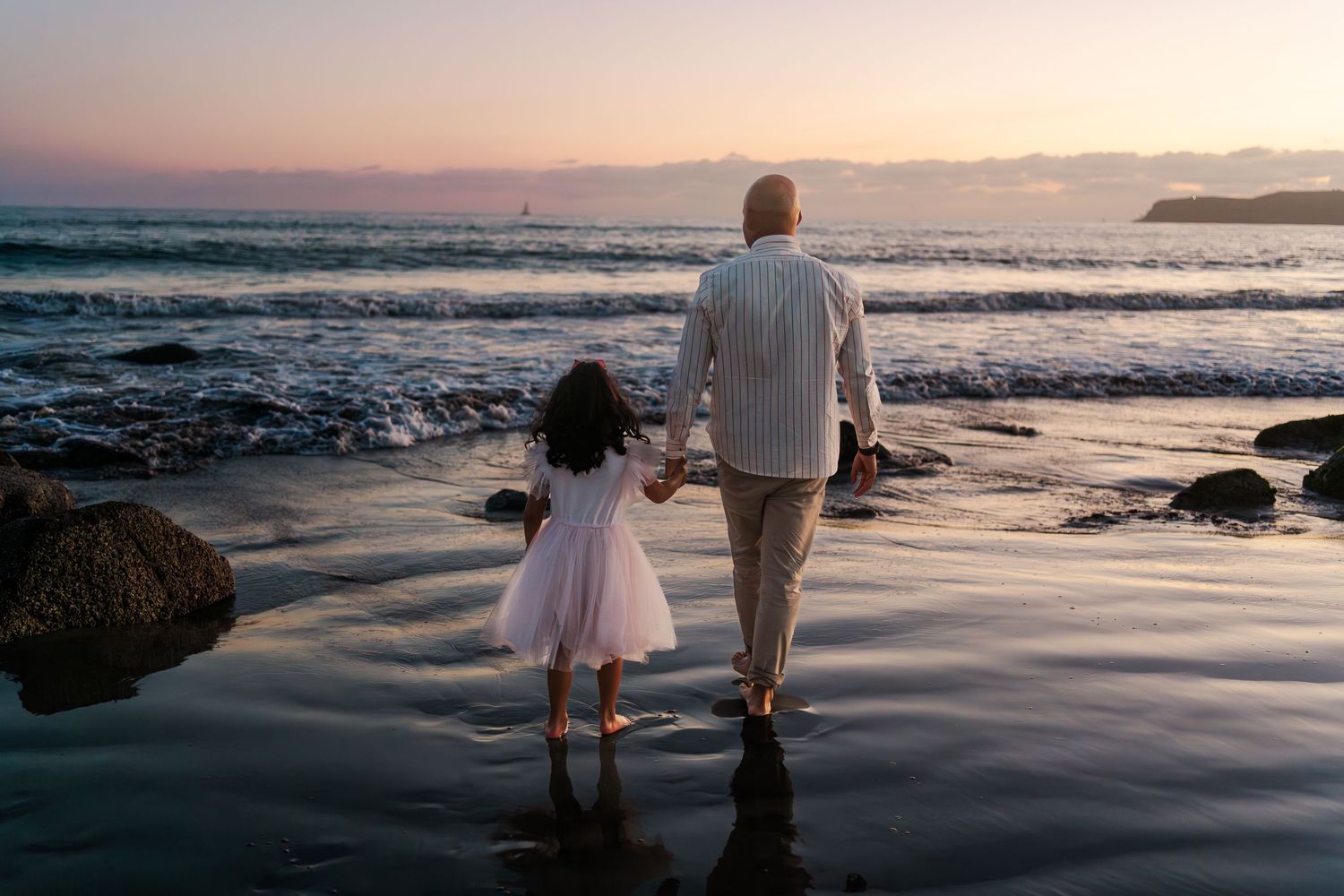 Figure in white dress and figure in striped shirt hold hands while walking on beach at sunset with waves crashing.