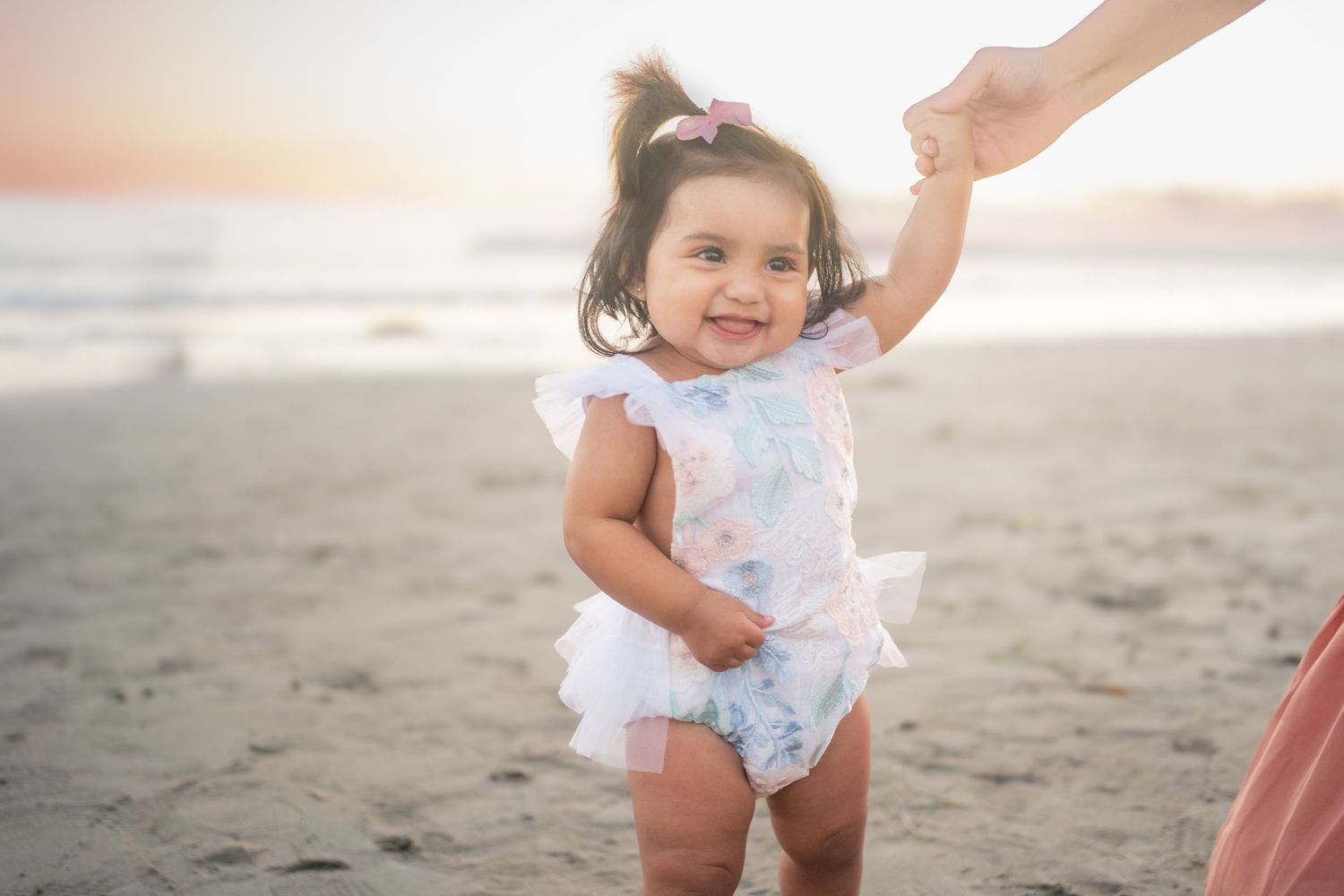 Small child wearing a floral romper holding an adult's hand while standing on a sunny beach at sunset.