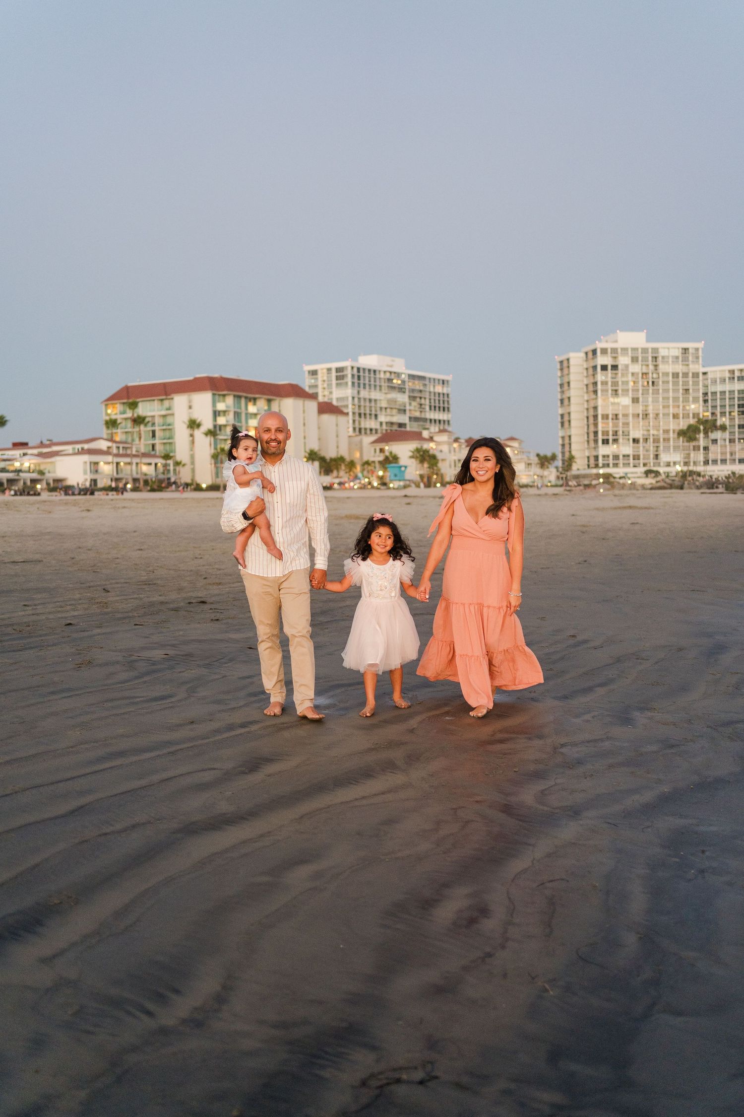 A family walks together on a beach at sunset with coastal buildings in the background.