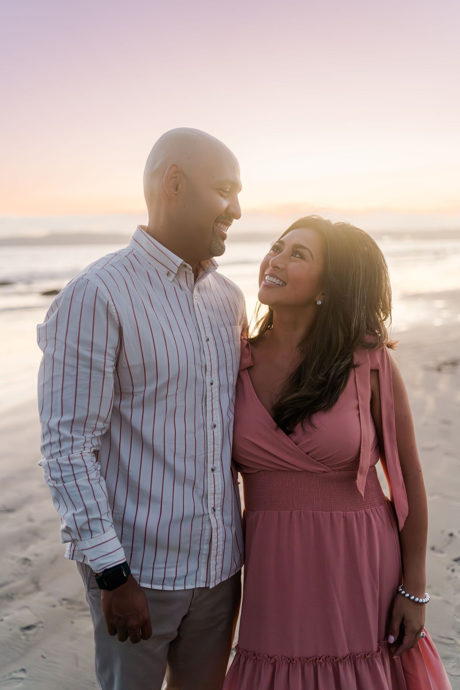 Happy couple sharing romantic moment together on beach at sunset wearing striped shirt and pink dress.