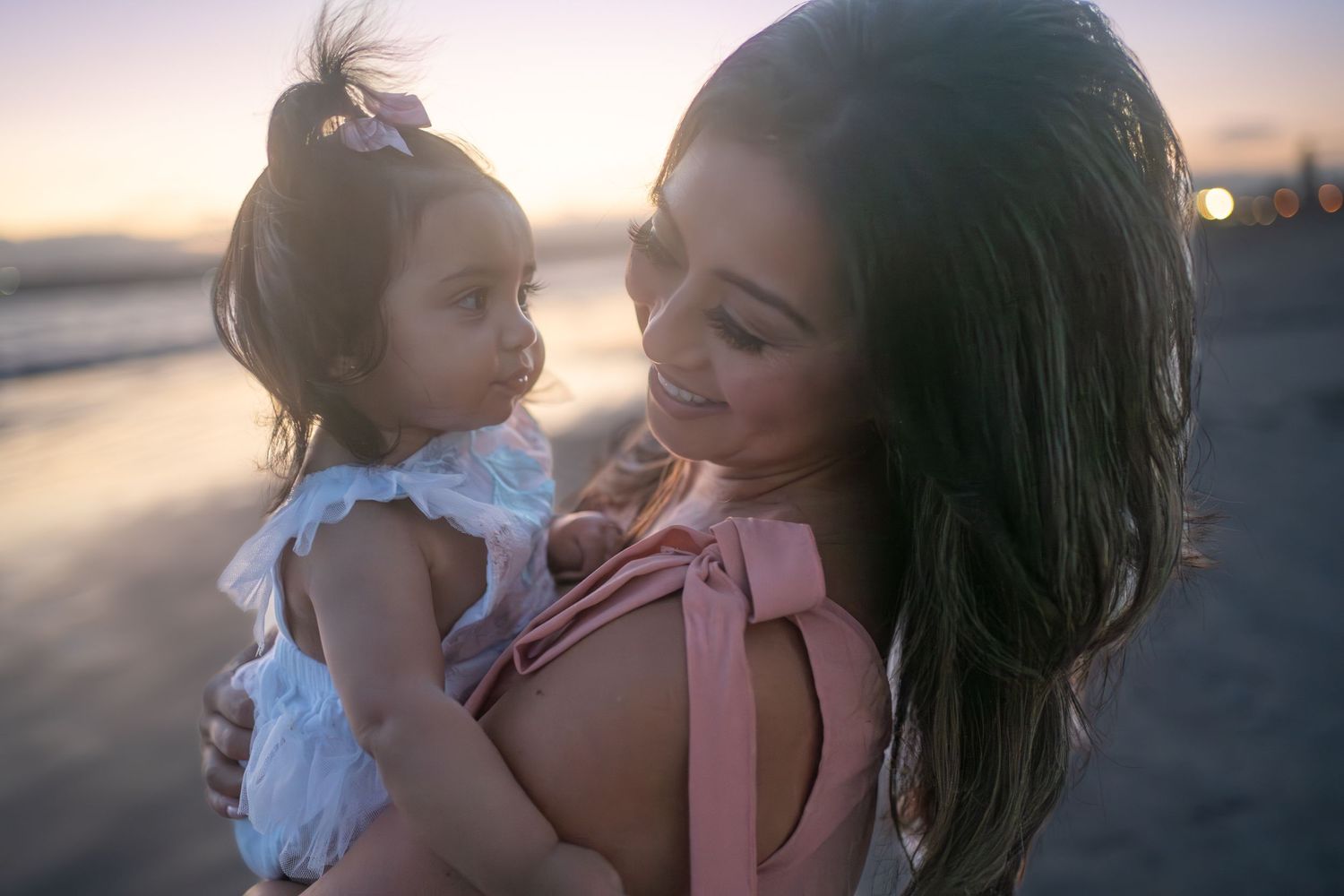 Mother and toddler share a tender moment by the beach at sunset, captured in golden light.
