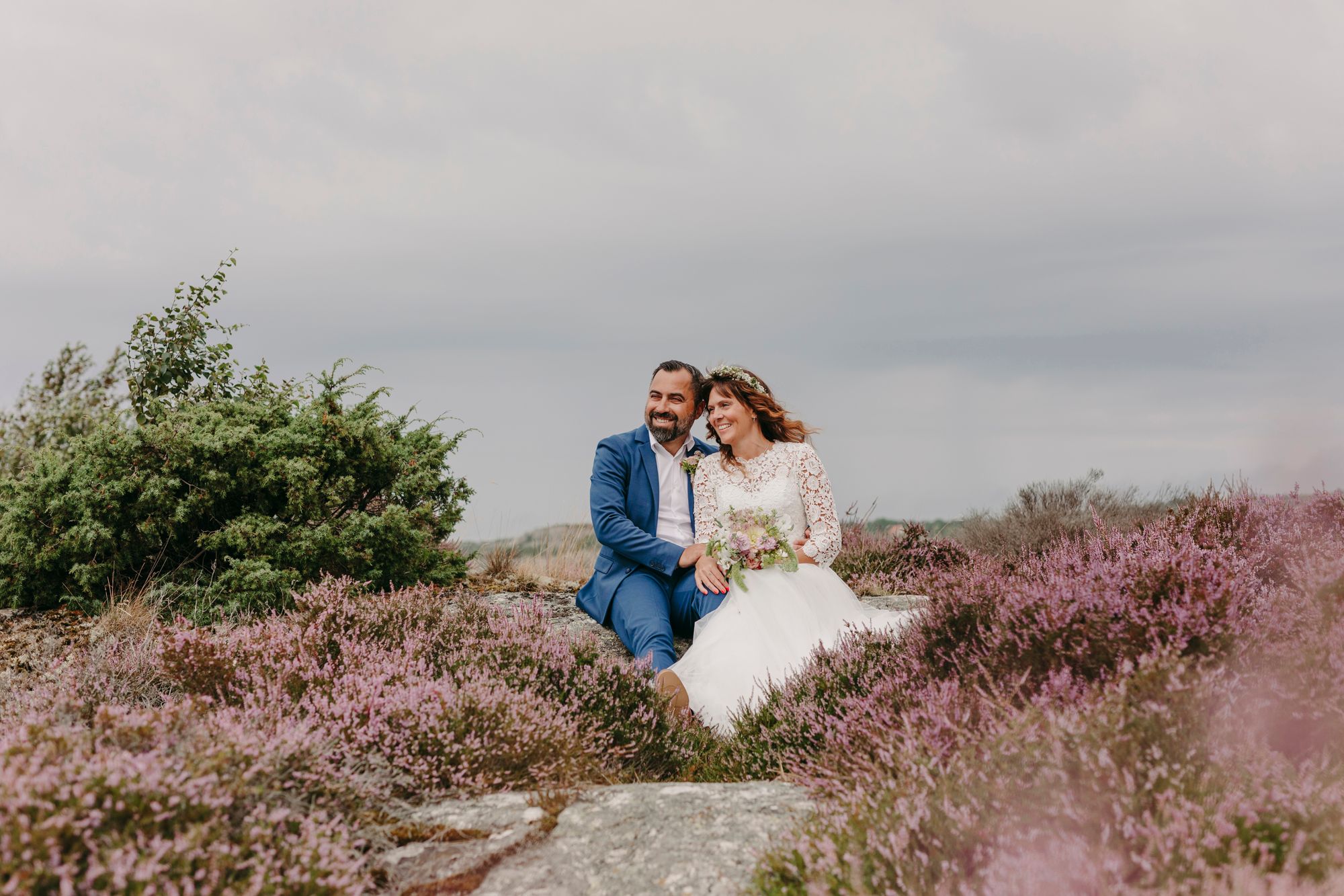 A couple shares a romantic moment surrounded by blooming purple heather on a cloudy hillside.