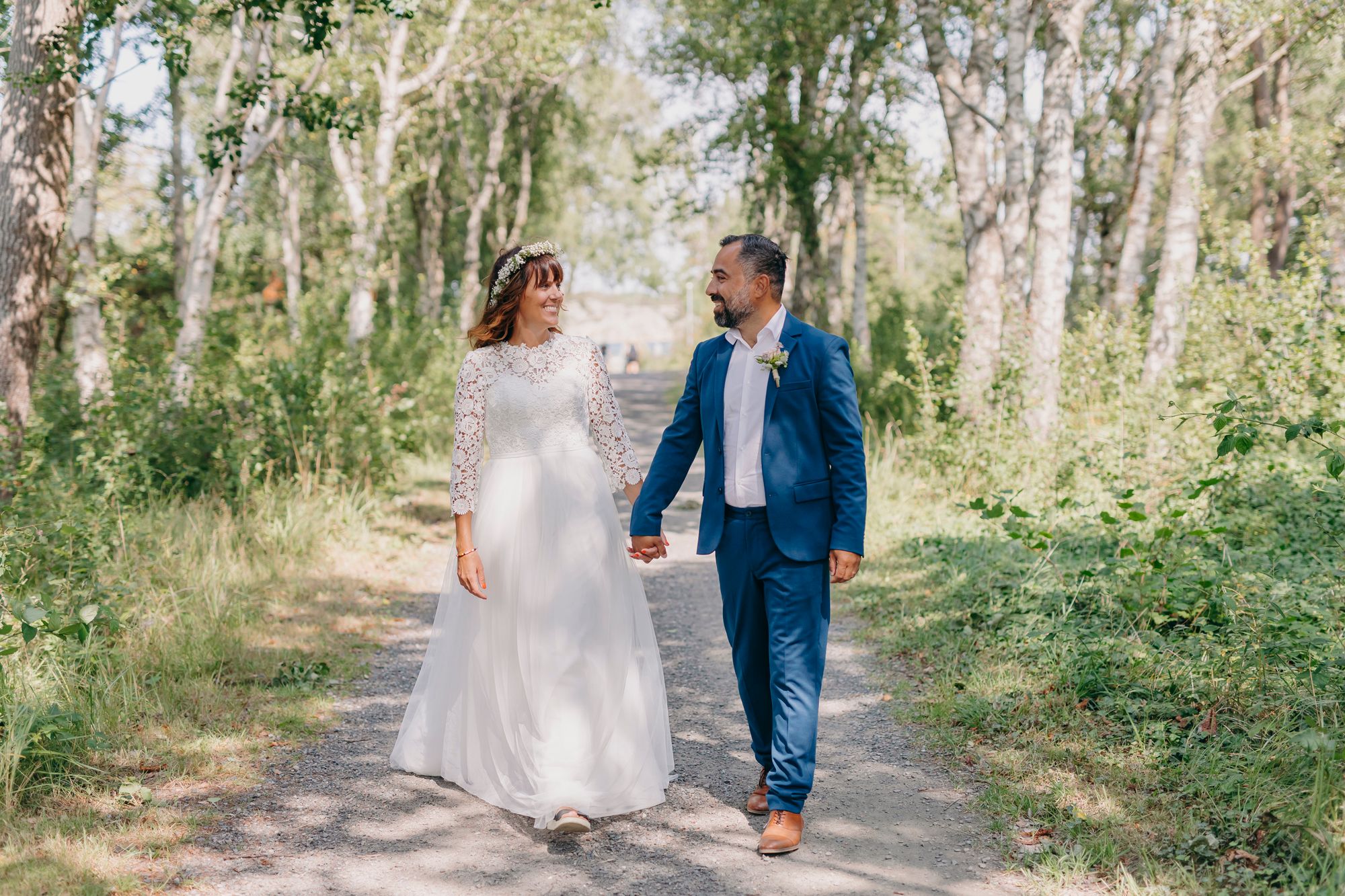 A couple walks hand in hand through a sunlit wooded path wearing wedding attire on their special day.