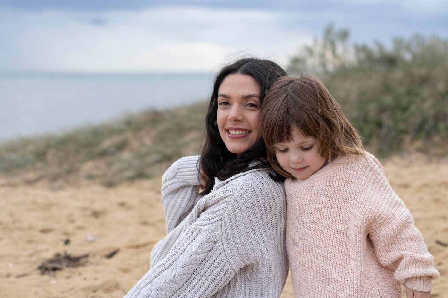 A parent and child in warm sweaters share a tender embrace on a cloudy beach.