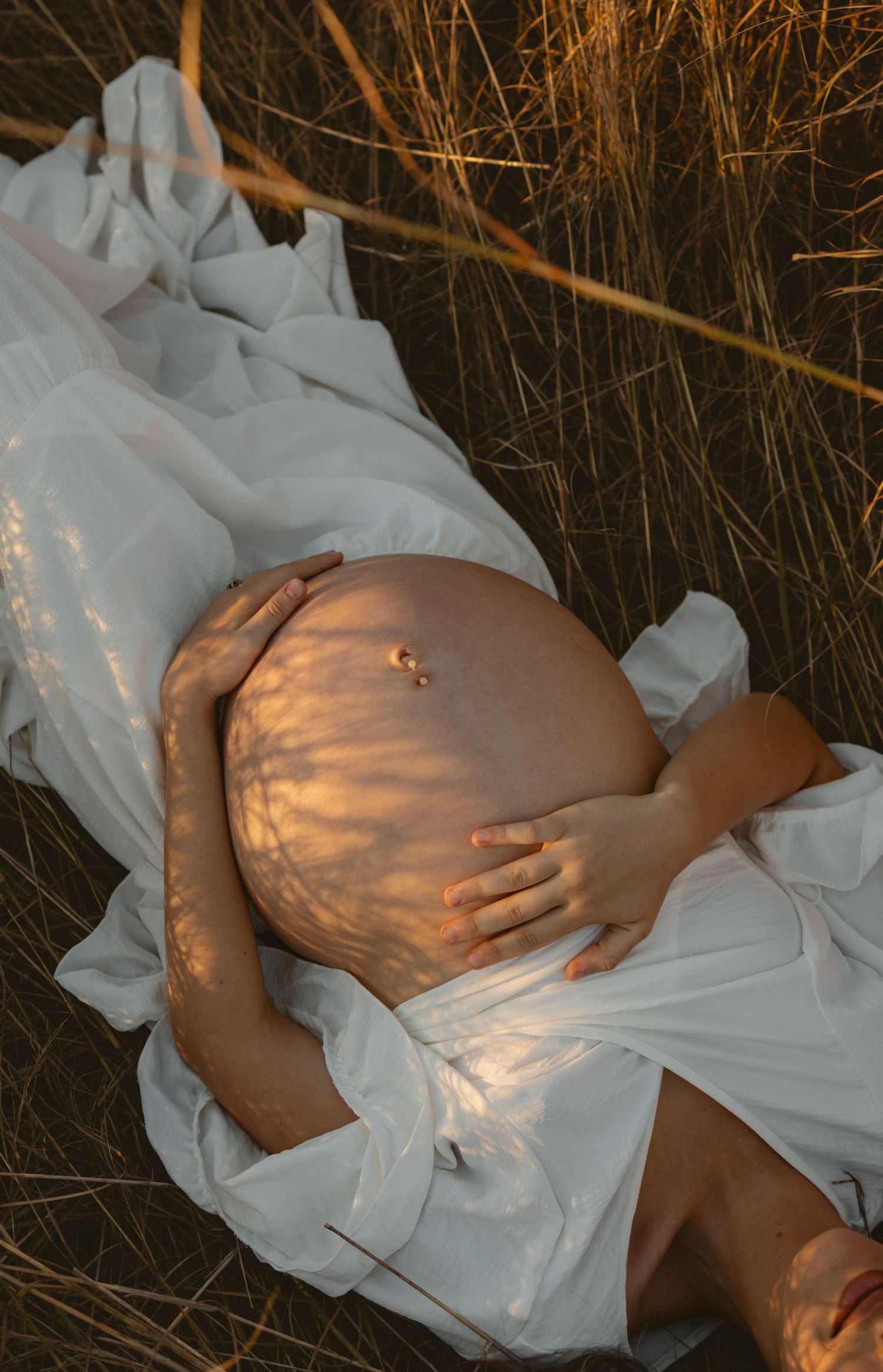 Artistic maternity photoshoot showing a pregnant belly illuminated by warm sunset light against tall grass and white fabric.