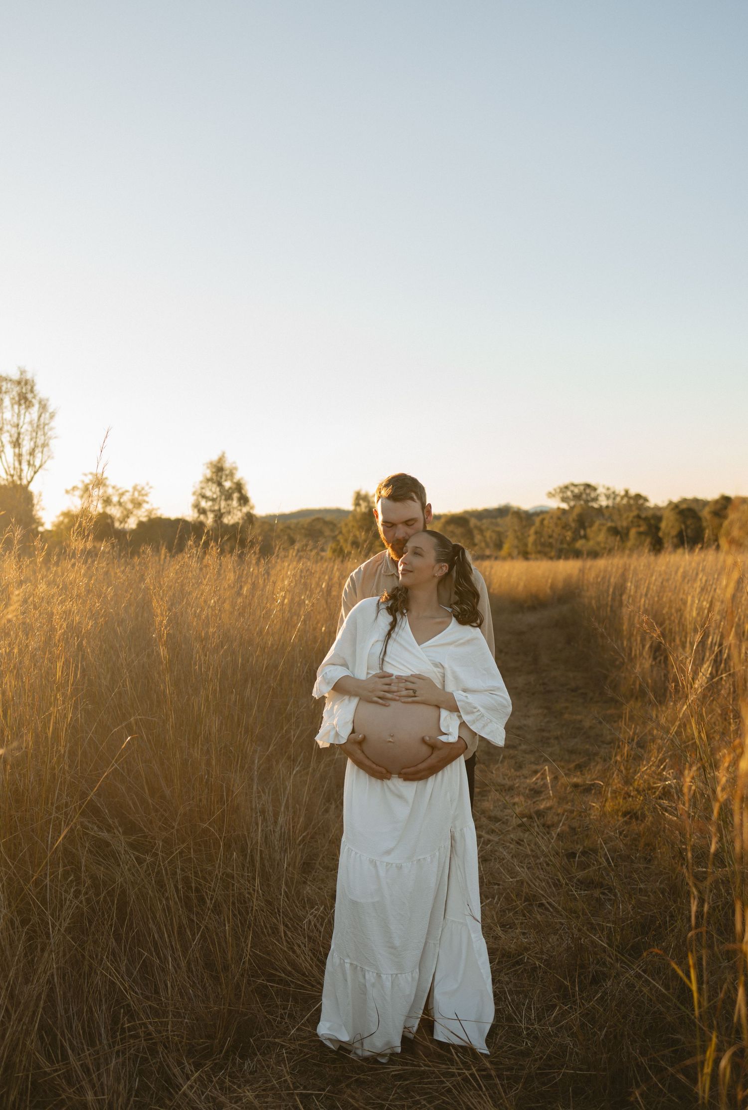 A couple in white clothing embraces tenderly in a sunlit meadow at golden hour.