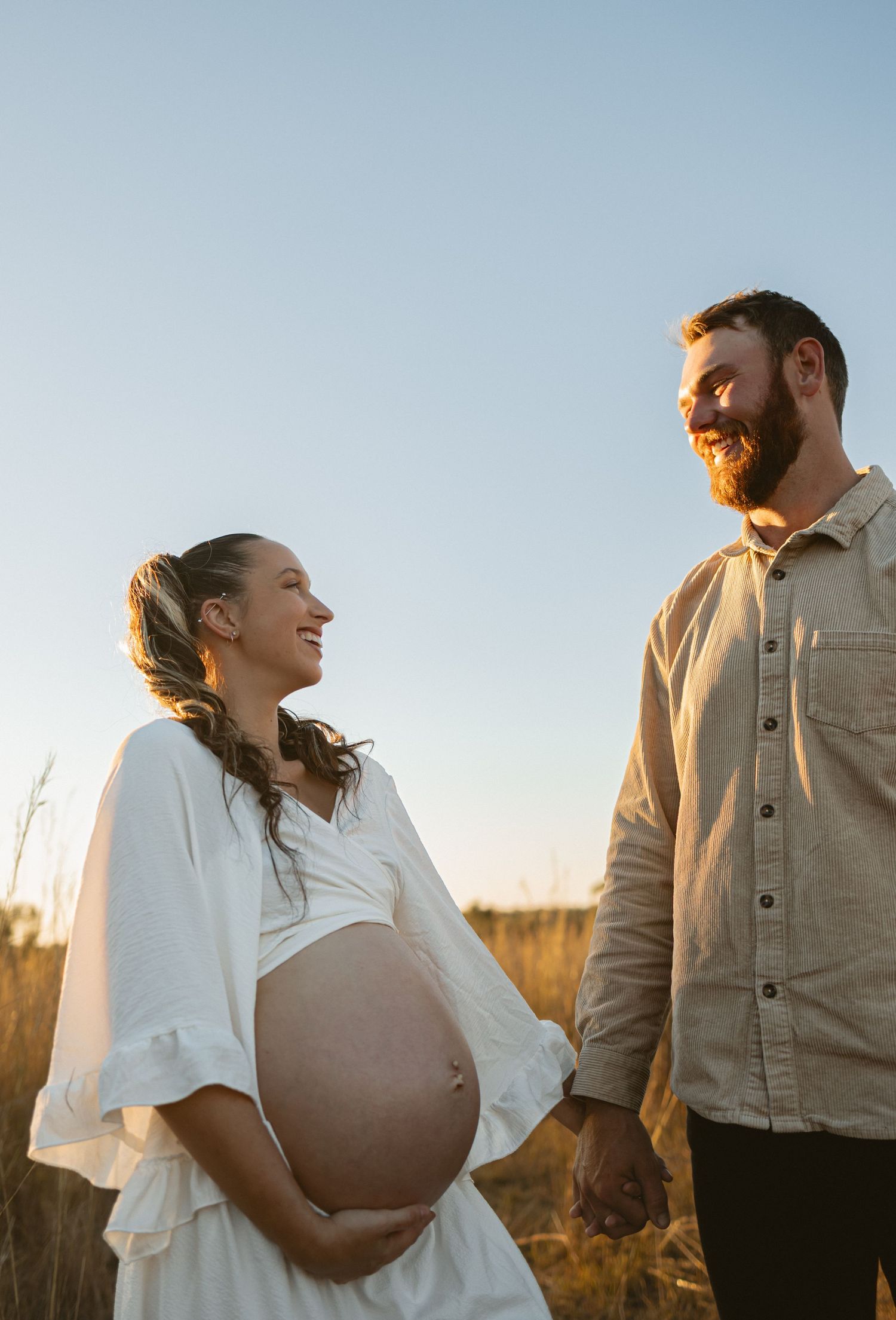 A couple embraces in a golden field during sunset while wearing white and tan outfits creating a romantic silhouette.