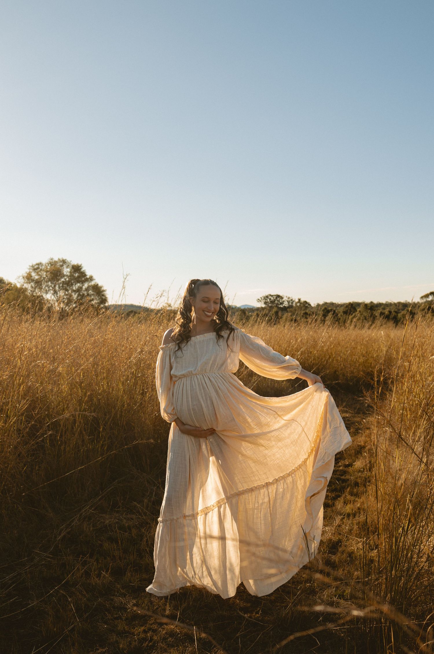 Figure in a flowing white dress stands ethereally in a golden field during sunset, creating a dreamlike silhouette.