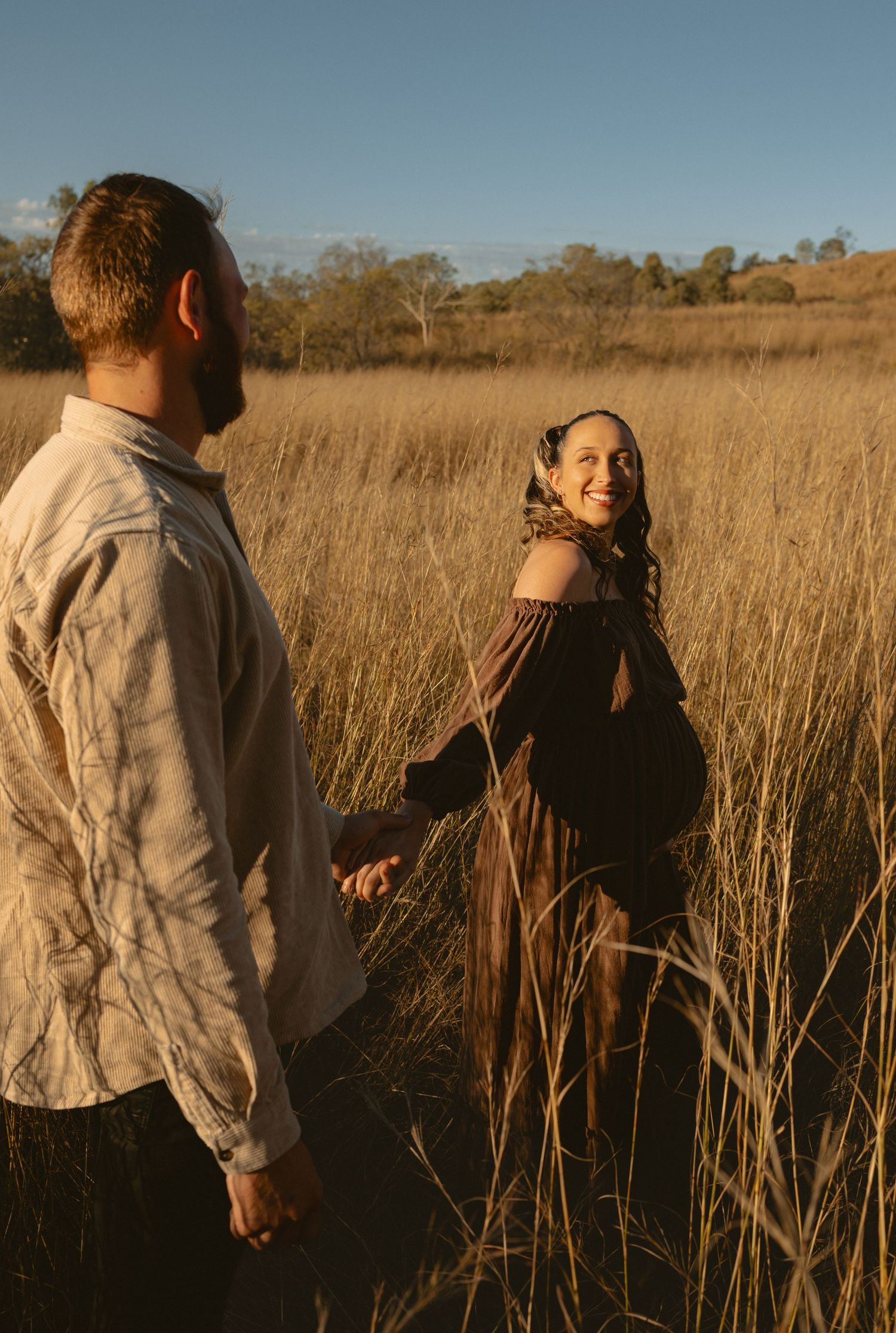 Two people in casual clothes walk through a golden field of tall wheat grass at sunset.