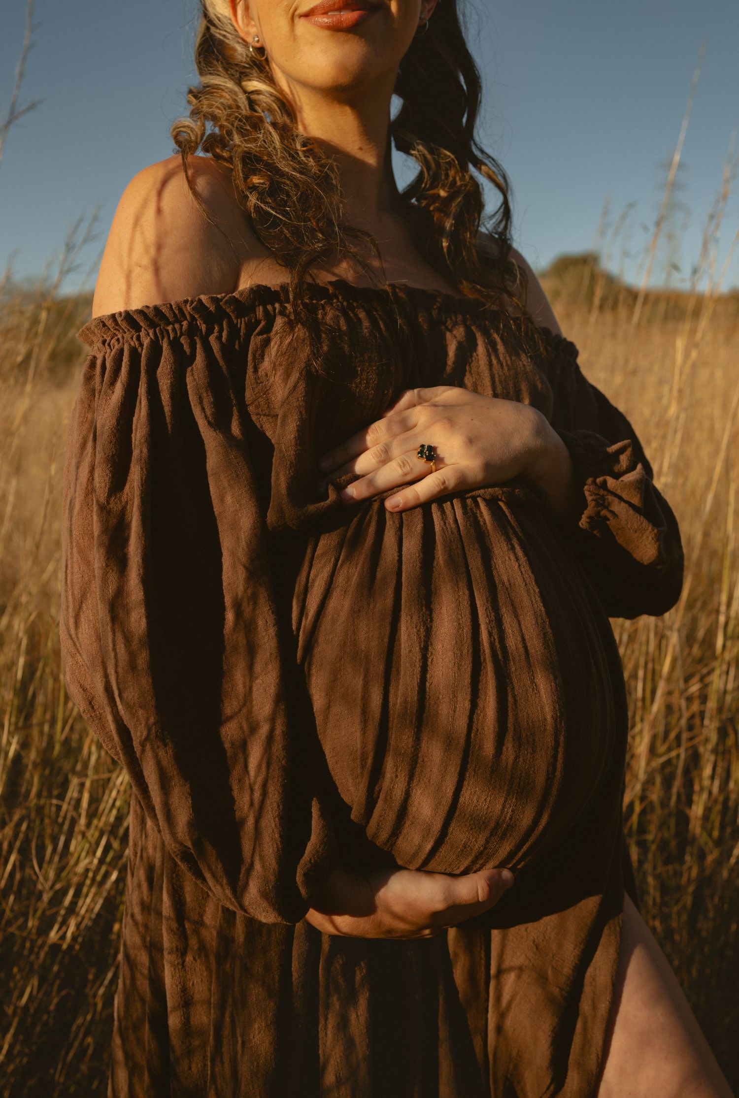 Someone in a brown flowy dress holds a small opossum in their arms while standing in a golden wheat field at sunset.