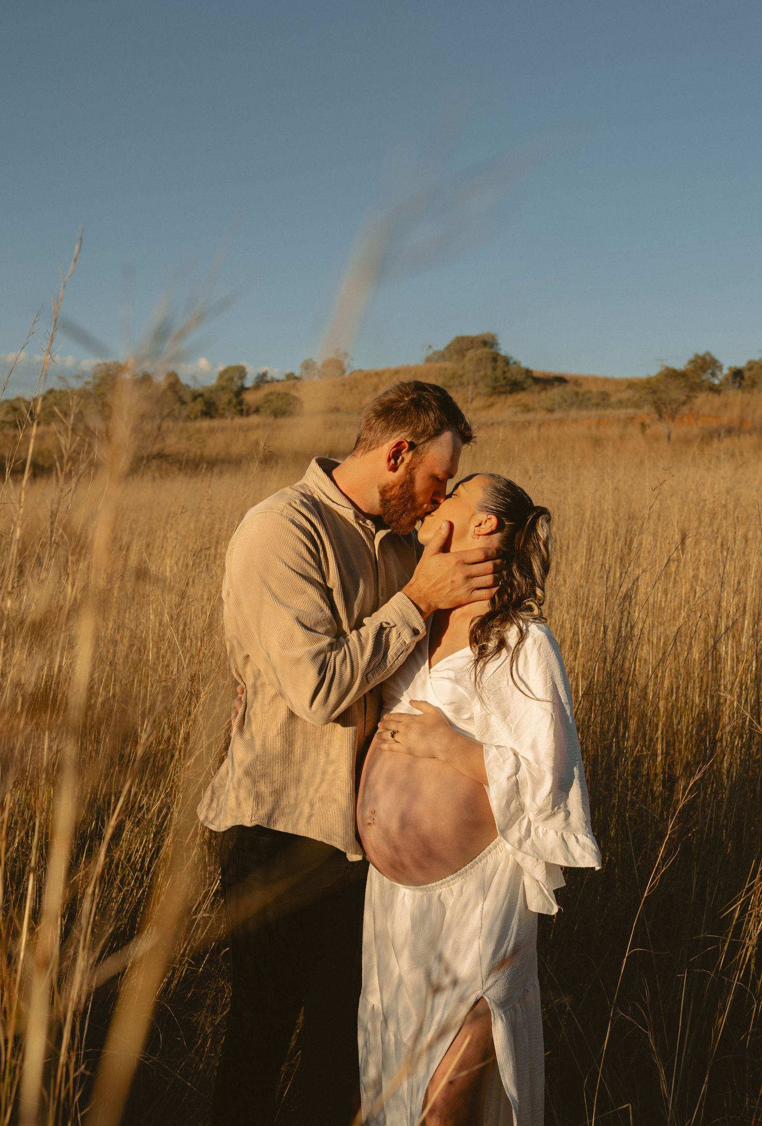 A romantic couple embraces and kisses in a golden field of tall grass during sunset.