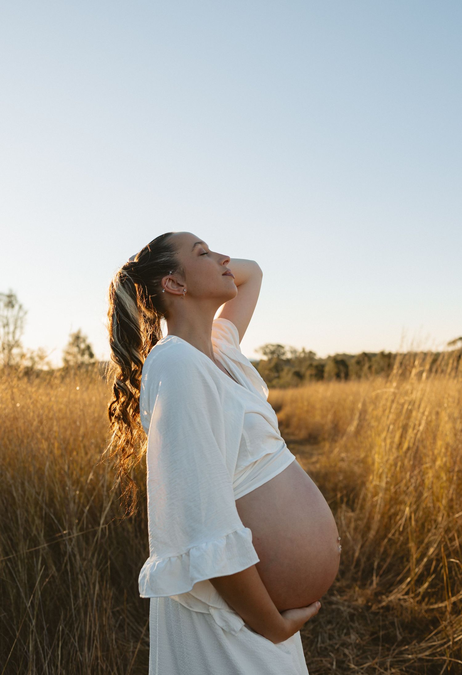 A pregnant woman stands in a golden field of tall grass during sunset wearing a white flowing dress.