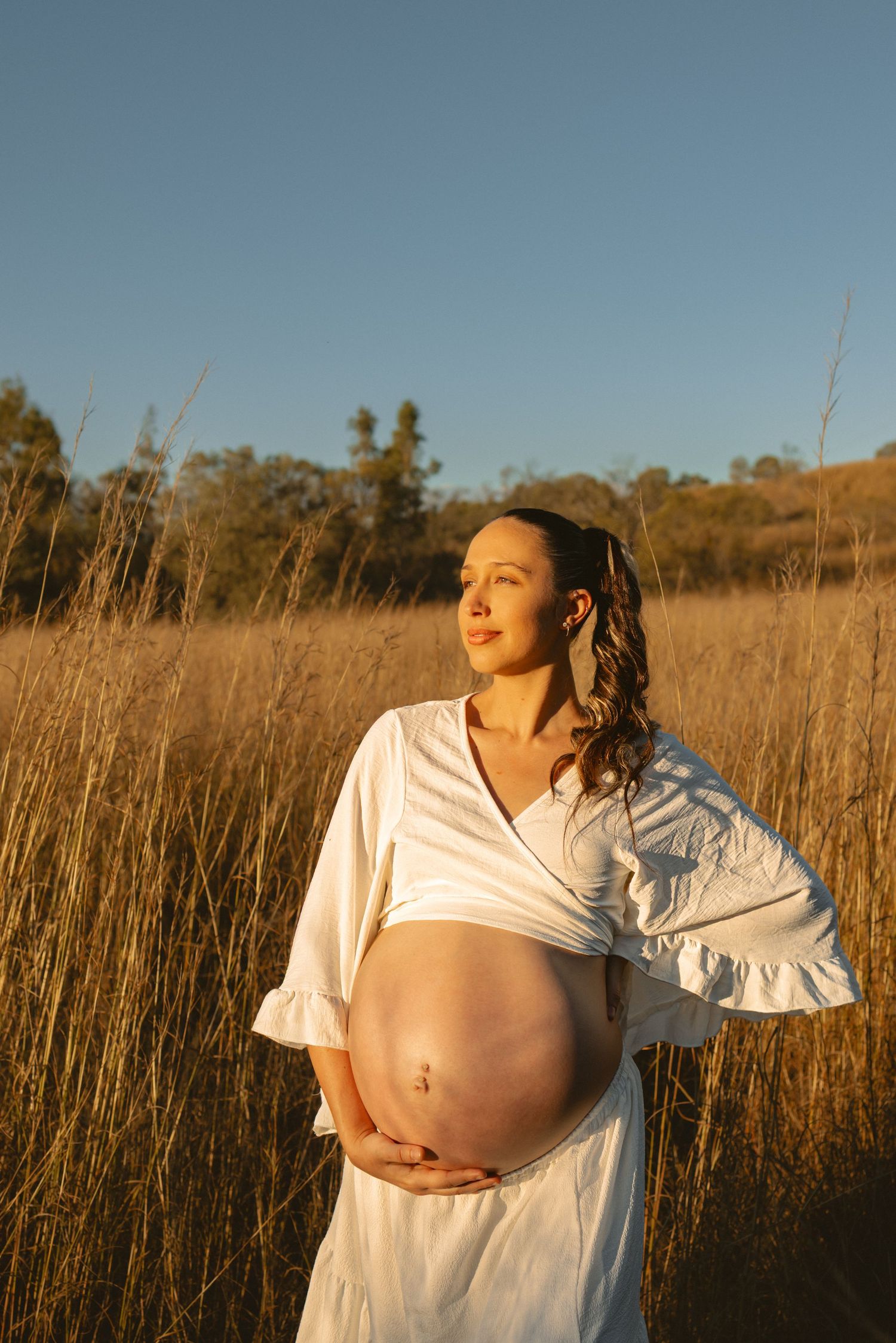 A maternity photo sequence shows a pregnant person in a white shirt standing in a golden field at sunset.