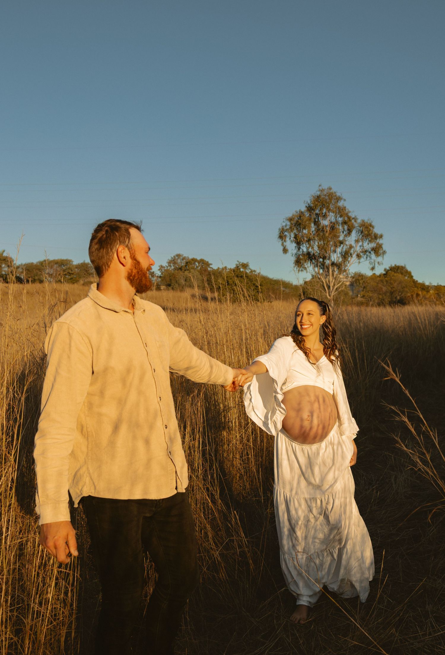 Two people walk together in tall grass during golden hour sunset in a dreamy outdoor setting.