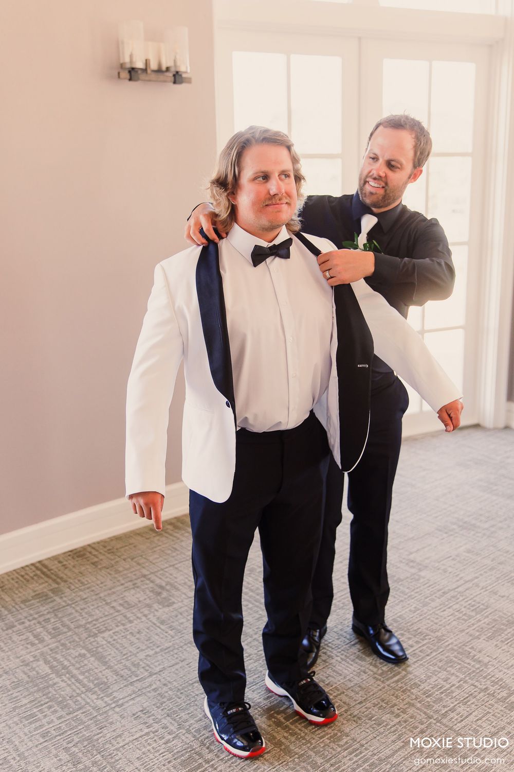 Two people in formalwear adjust a bowtie and suspenders before a wedding celebration.