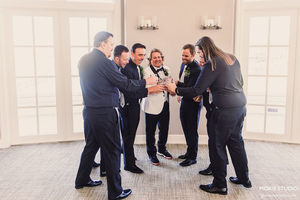 A group of people in dark formal attire practice dance moves together in a bright sunlit studio space.