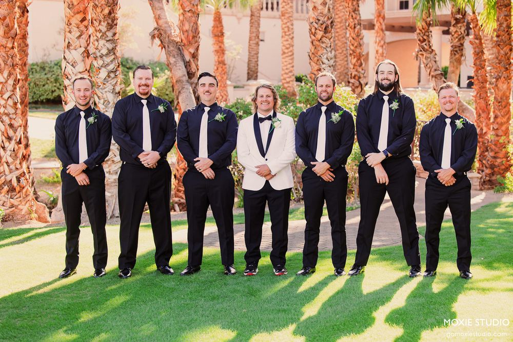 A wedding party in black tuxedos poses in a line against a backdrop of palm trees and coral-colored columns.